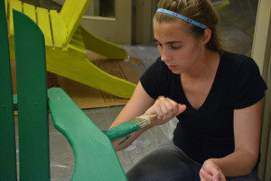 A young woman applies green paint to the arm of a wooden Adirondack chair.